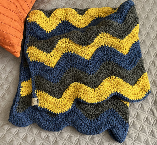 Crocheted blanket with blue, yellow, and gray chevron pattern on a textured surface.